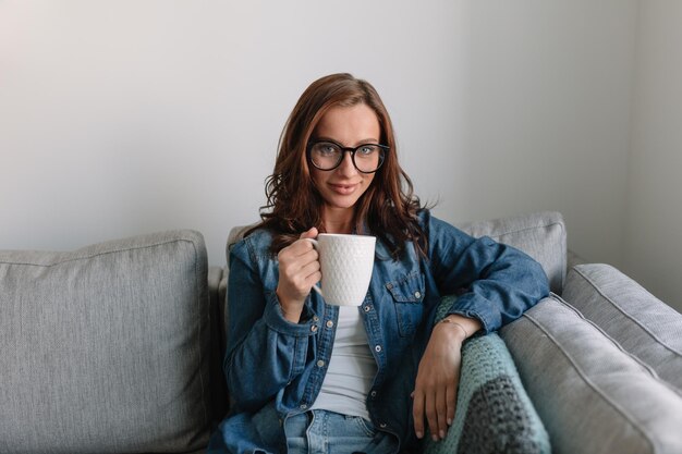 Portrait intérieur d'une fille caucasienne avec un sourire blanc comme neige regarde dans l'appareil photo sur fond blanc à la maison beauté brune dans des lunettes rondes t-shirt blanc et chemisier