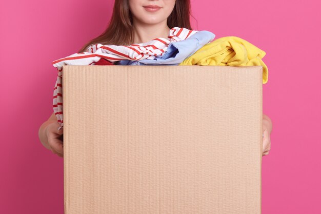 Portrait intérieur d'une fille sans visage debout avec une boîte en carton dans les mains, tenant une boîte en carton pleine de vêtements à la mode isolés sur un mur rose. Concept de don, de charité et de bénévolat.