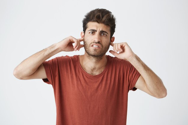 Portrait intérieur d'un gars mature espagnol en colère portant un t-shirt rouge à la colère et bouchant les oreilles avec les doigts en dispute avec sa petite amie.