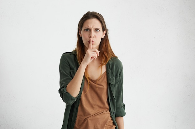 Portrait intérieur isolé de la jeune femme européenne en colère folle dans des vêtements décontractés fronçant les sourcils, faisant des gestes avec le doigt, le gardant sur les lèvres, demandant de garder le silence