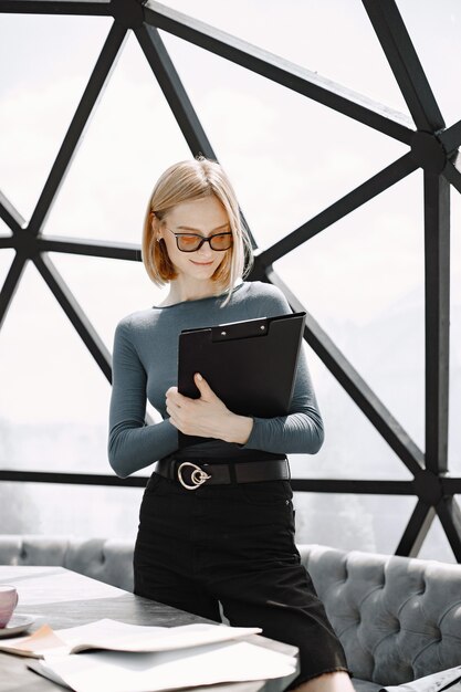 Portrait intérieur d'une jeune femme d'affaires debout dans un café et écrivant sur un porte-papier. Fille blonde portant des lunettes de soleil