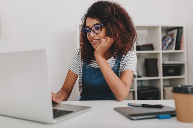 Portrait intérieur de jeune femme noire dans des verres surfer sur internet assis devant des étagères avec des documents