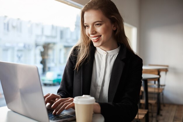 Portrait intérieur de jolie femme européenne assise dans un café, boire du café et taper dans un ordinateur portable, être heureux et heureux.