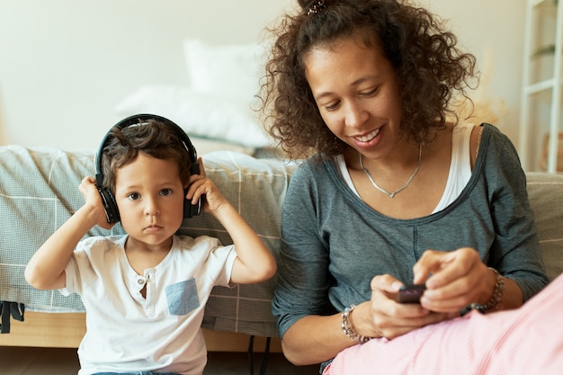 Portrait intérieur de joyeuse jeune femme hispanique tenant un téléphone mobile jouant des morceaux de musique pour son adorable petit fils qui écoute des chansons via des écouteurs sans fil