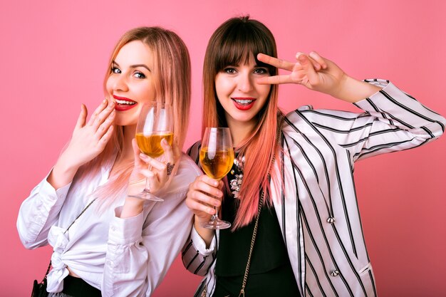 Portrait intérieur positif de deux jolies femmes élégantes et élégantes s'amusant à la fête, buvant du champagne savoureux et dansant, tenues de soirée cocktail et mur rose