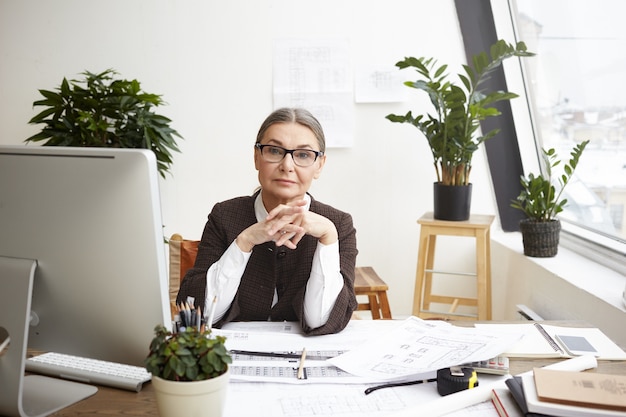 Portrait à l'intérieur d'un professionnel sérieux 55 ans femme architecte senior étudiant les plans architecturaux, vérification des mesures sur ordinateur et correction des dessins sur 24 en face d'elle