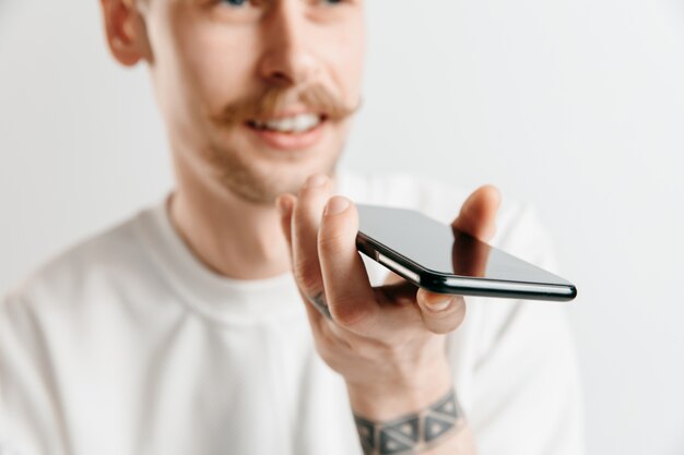 Portrait intérieur de séduisant jeune homme isolé sur un espace gris, tenant un smartphone, à l'aide de la commande vocale, se sentir heureux et surpris