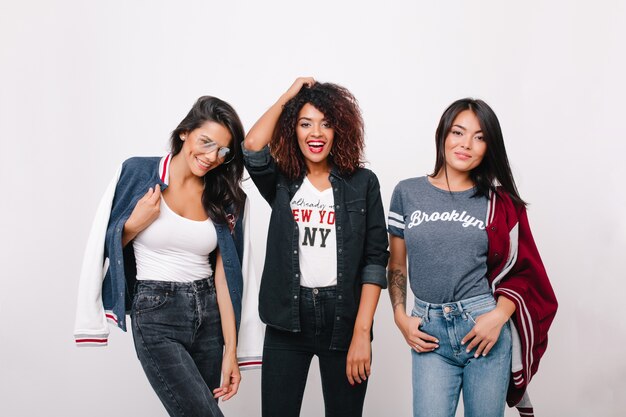 Portrait intérieur de trois étudiantes excitées dans des vêtements à la mode s'amusant ensemble après les cours. Fille bouclée en tenue de denim, passer du temps avec des amis brune et rire.