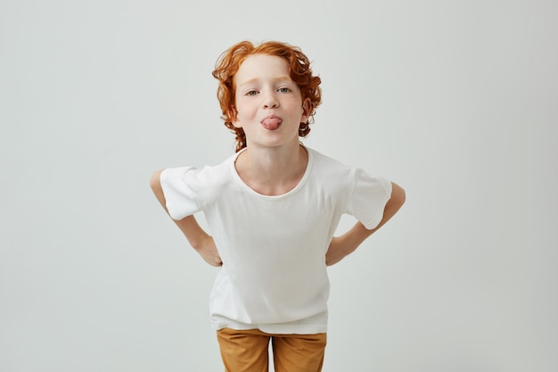 Portrait isolé de l'adorable petit garçon drôle avec des cheveux roux tenant les mains sur la taille, s'amusant et montrant la langue