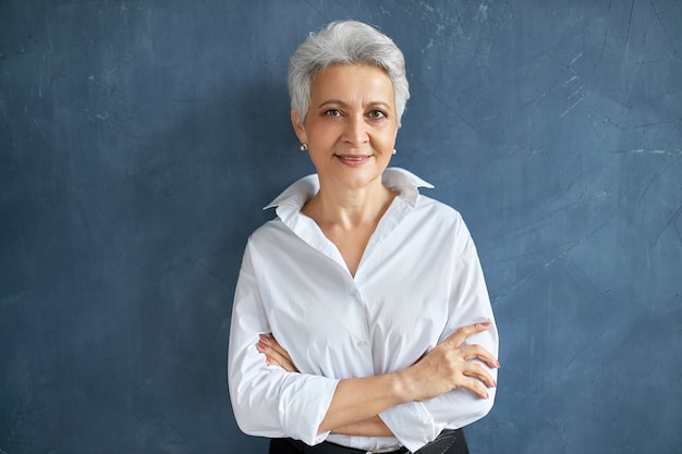 Portrait isolé de l'élégant courtier femme de 50 ans en chemise blanche posant sur un mur blanc