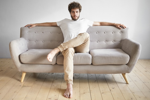 Portrait isolé de jeune homme européen à la mode à la mode avec une barbe épaisse se reposer à la maison, assis avec désinvolture sur un canapé luxueux, regarder la télévision, se sentir détendu. Gens, style de vie et loisirs
