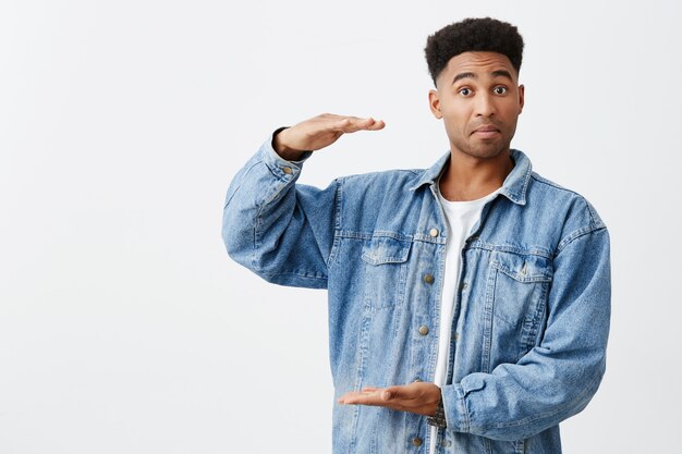 Portrait isolé de jeune homme à la peau foncée drôle avec une coiffure afro en chemise blanche décontractée sous une veste en jean faisant semblant de tenir une grande boîte en mains avec une expression de visage excité