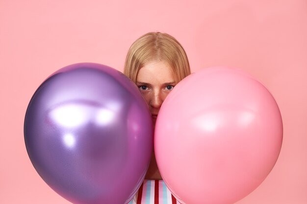 Portrait isolé de mystérieuse jeune femme blonde avec des taches de rousseur et piercing facial posant sur rose se cachant derrière deux ballons d'hélium métallique brillant