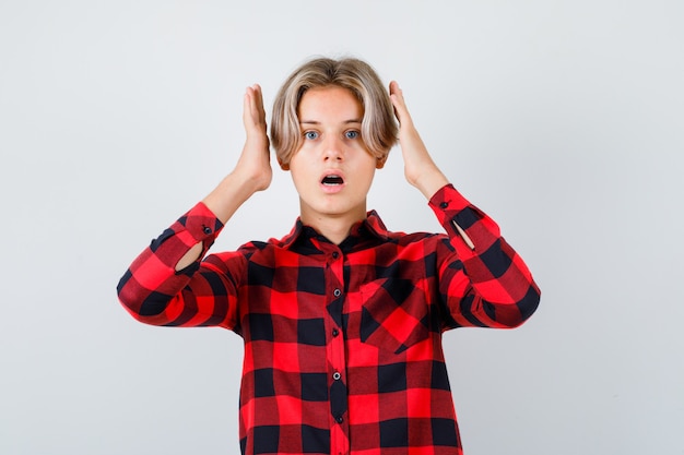 Portrait de jeune adolescent avec les mains près de la tête en chemise à carreaux et à la vue de face choquée