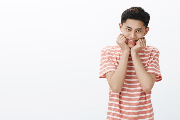 Portrait de jeune adolescent mâle asiatique idiot et mignon en t-shirt rayé souriant tête penchée sur les paumes pressées à mâchoire essayant de regarder doux et tendre