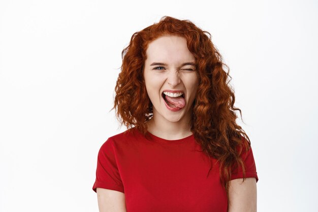 Portrait d'une jeune adolescente au gingembre insouciante faisant des grimaces positives, montrant la langue et souriant, un clin d'œil pour vous motiver et vous encourager, mur blanc