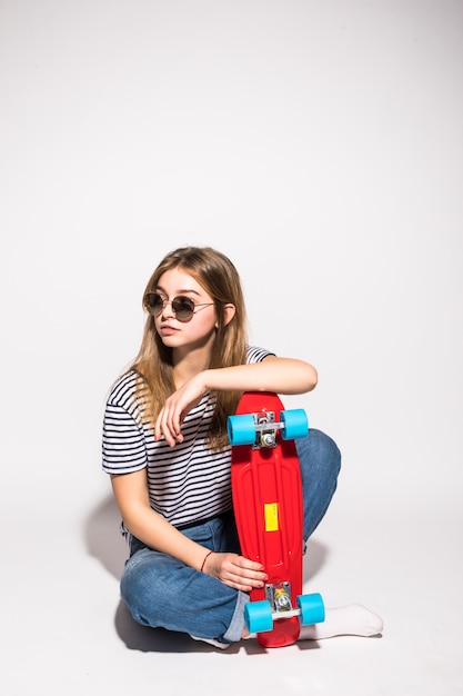 Portrait de jeune adolescente à lunettes posant avec planche à roulettes en se tenant debout sur le mur blanc