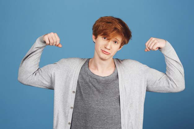 Portrait de jeune beau mec aux cheveux roux dans des vêtements gris décontractés jouant des muscles, avec une expression incertaine et confuse, se sentant mal à l'aise.