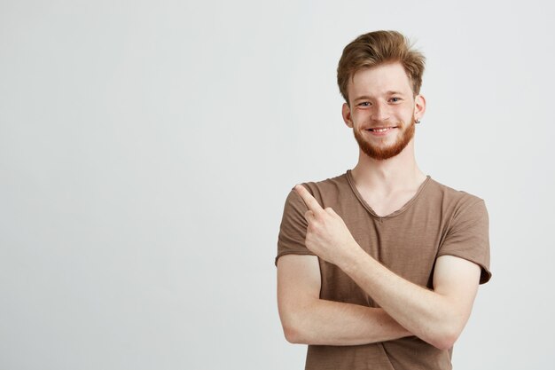 Portrait de jeune bel homme sincère avec barbe souriant pointant le doigt sur le côté.