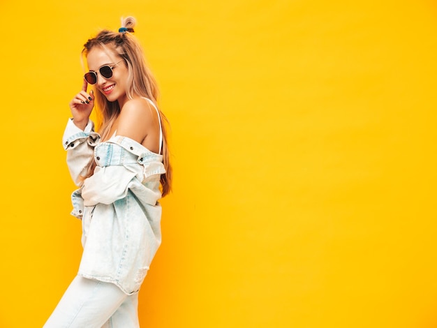 Portrait de jeune belle femme blonde souriante dans des vêtements d'été à la mode Sexy femme insouciante posant près du mur jaune en studio Modèle positif s'amusant à l'intérieur Gai et heureux en lunettes de soleil