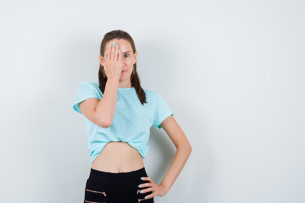 Portrait de jeune belle femme couvrant les yeux avec la main en t-shirt, pantalon et regardant la vue de face curieuse