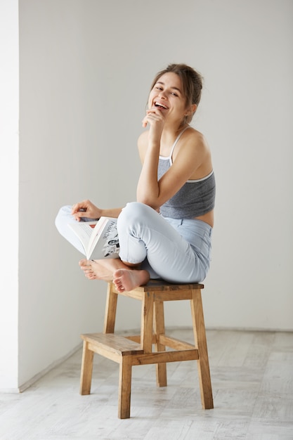 Portrait de jeune belle femme heureuse souriant rire tenant un livre assis sur une chaise sur un mur blanc à la maison.