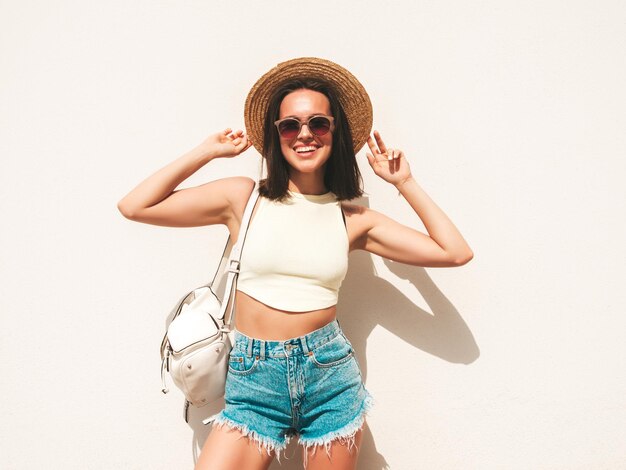 Portrait de jeune belle femme hipster souriante en short et chapeau de jeans d'été à la mode Modèle insouciant sexy posant dans la rue près du mur blanc Modèle positif à l'extérieur avec une malletteMontre la langue