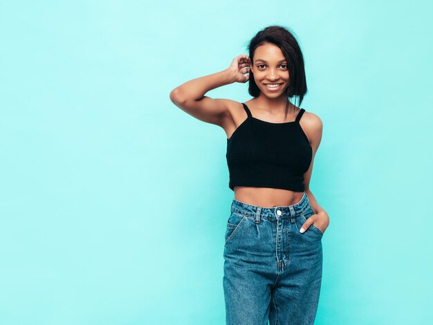 Portrait de jeune belle femme noire Modèle souriant vêtu d'un jean d'été et de hauts vêtements noirs Sexy femme insouciante posant près du mur bleu en studio Bronzé et joyeux