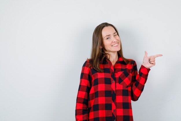 Portrait de jeune belle femme pointant vers la droite, un clin d'œil en chemise décontractée et à la vue de face joyeuse