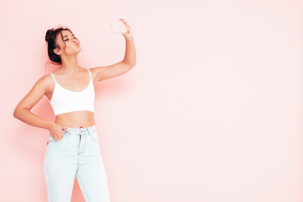 Portrait de jeune belle femme souriante dans des vêtements d'été à la mode Sexy femme insouciante posant près d'un mur rose en studio Modèle positif s'amusant à l'intérieur Prendre des photos de selfie