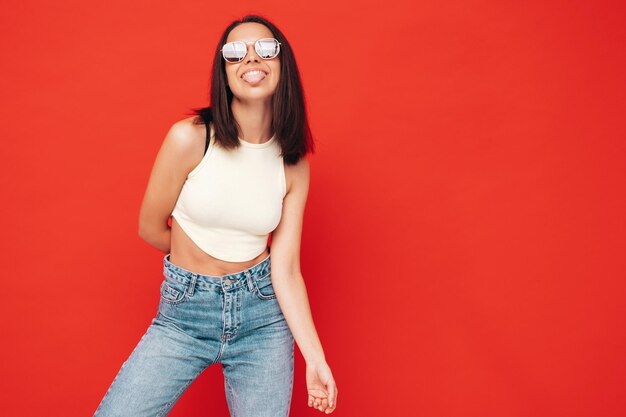 Portrait de jeune belle femme souriante dans des vêtements d'été à la mode Sexy femme insouciante posant près d'un mur rouge en studio Modèle positif s'amusant à l'intérieur Dans des lunettes de soleil Montre la langue