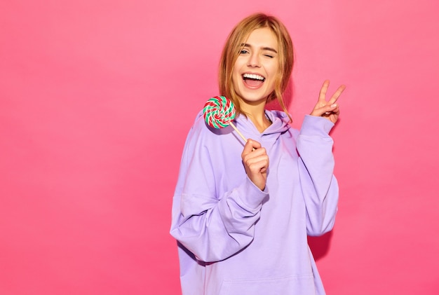 Portrait de jeune belle femme souriante hipster en hoodie d'été à la mode. Sexy femme insouciante posant près du mur rose. Modèle positif avec sucette montrant le signe de la paix