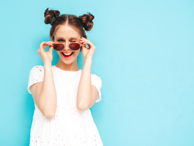 Portrait de jeune belle femme souriante en robe blanche d'été à la mode femme insouciante posant près du mur bleu en studio Modèle positif s'amusant à l'intérieur Enthousiaste et heureux Enlève ses lunettes de soleil