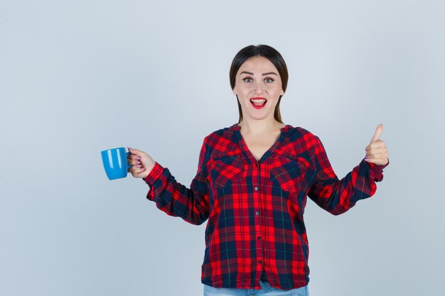 Portrait De Jeune Belle Femme Tenant Une Tasse Tout En Montrant Le Pouce Vers Le Haut En Chemise Décontractée, En Jean Et En Regardant La Vue De Face Joyeuse