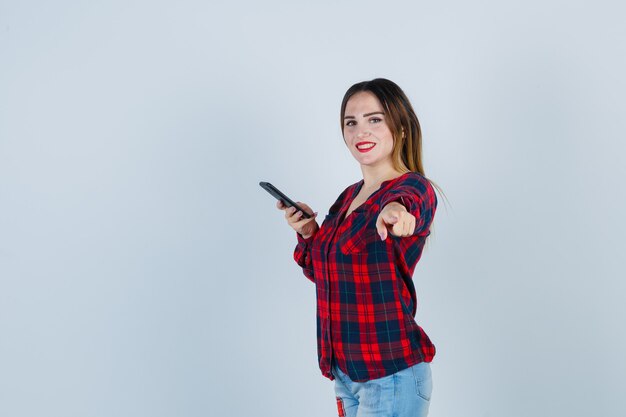Portrait de jeune belle femme tenant un téléphone, pointant vers l'avant en chemise décontractée et à la vue de face heureuse