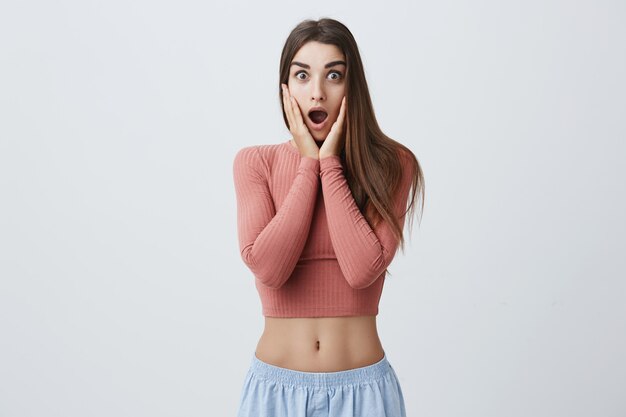 Portrait de jeune belle fille étudiante drôle caucasien aux longs cheveux bruns en tenue à la mode serrant le visage avec les mains avec la bouche ouverte