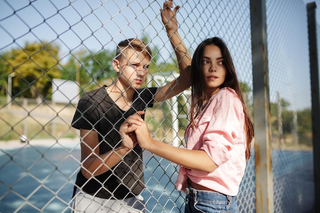 Portrait de jeune belle fille et garçon debout entre une clôture en maille sur un terrain de basket et regardant pensivement de côté