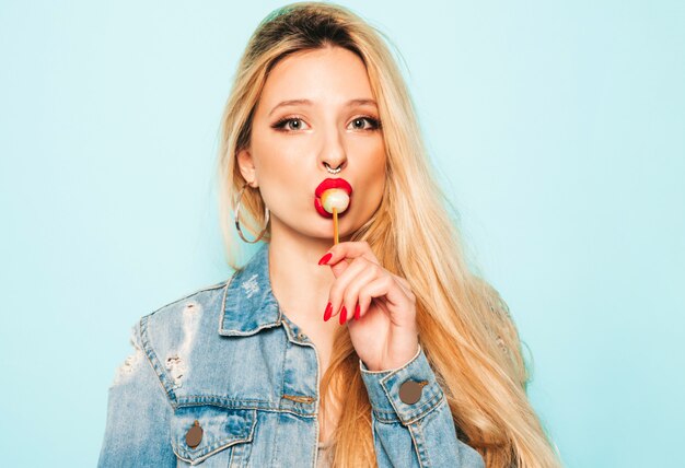 Portrait de jeune belle fille mauvaise hipster dans des vêtements jeans à la mode et boucle d'oreille dans son nez. Modèle positif léchant des bonbons de sucre ronds