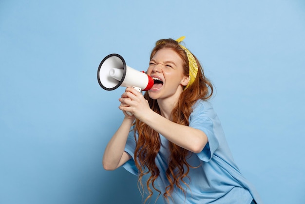 Portrait de jeune belle fille rousse criant dans un mégaphone isolé sur fond de studio bleu