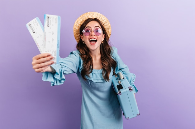 Photo gratuite portrait de jeune brune au chapeau et lunettes posant avec valise sur mur violet. femme en robe bleue se réjouissant sincèrement de voyager.