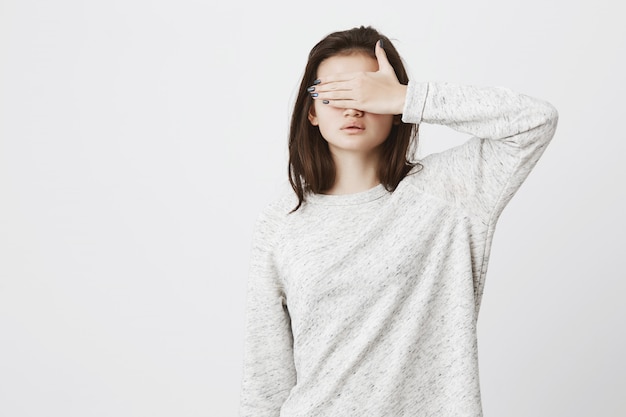Portrait de jeune brune mignonne dans des vêtements à la mode, couvrant ses yeux avec la main, debout sur un mur blanc.