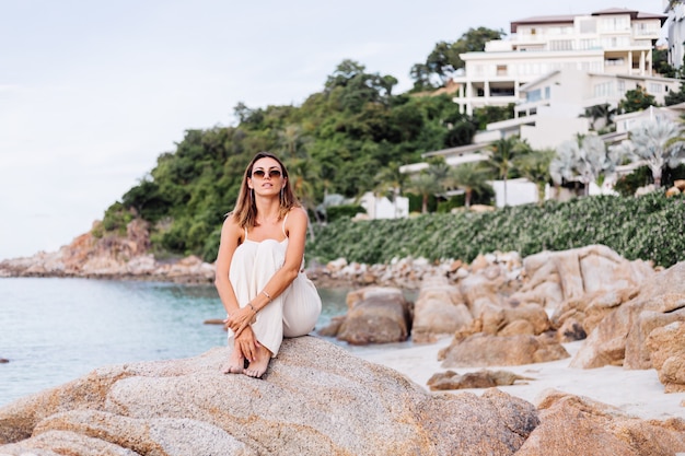 Portrait de jeune calme heureux caucasien fit slim woman in crop cami top et ensemble de pantalons se trouve seul sur une plage tropicale rocheuse au coucher du soleil