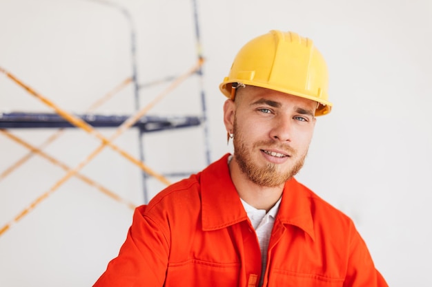 Photo gratuite portrait de jeune constructeur en vêtements de travail orange et casque jaune avec un crayon derrière l'oreille regardant rêveusement à huis clos avec des échafaudages sur fond