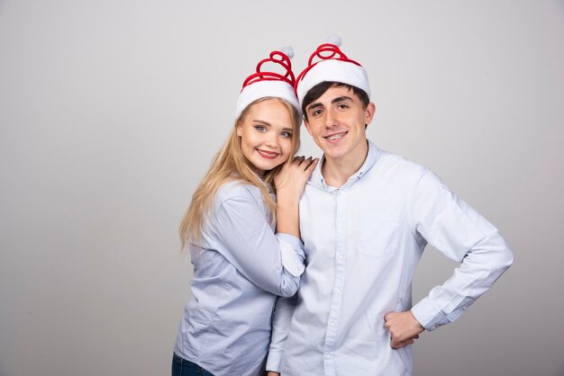 Portrait de jeune couple en bonnet de Noel posant joyeusement sur un mur gris.