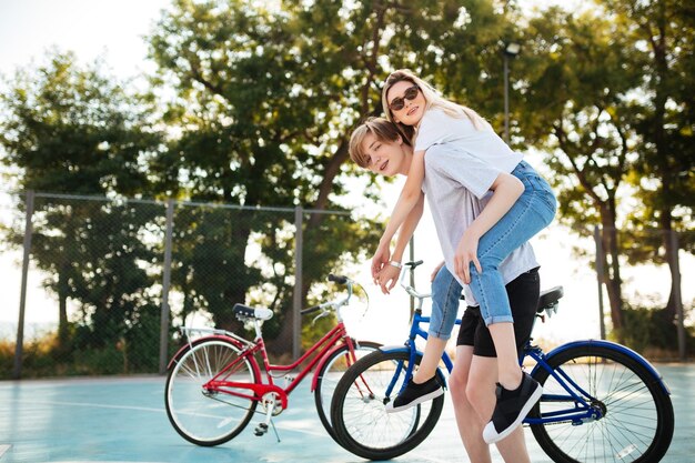 Portrait de jeune couple s'amusant dans le parc avec vélo rouge et bleu sur fond Garçon souriant regardant à huis clos tout en jouant avec une belle fille à lunettes de soleil et la tenant sur son dos