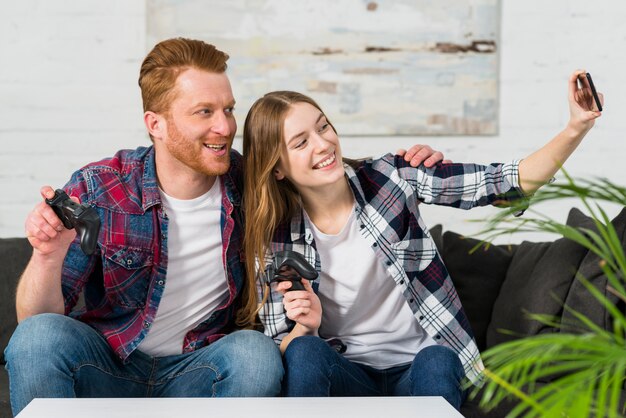 Photo gratuite portrait d'un jeune couple souriant tenant le contrôleur de jeu vidéo prenant selfie sur téléphone mobile