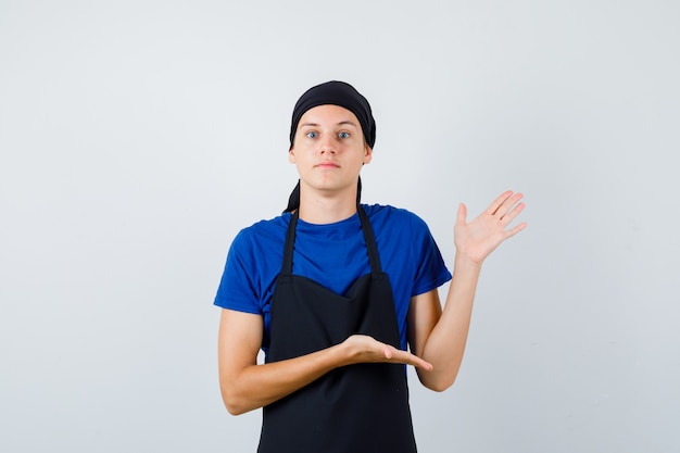 Portrait De Jeune Cuisinier Adolescent Faisant Semblant De Montrer Quelque Chose En T-shirt, Tablier Et à La Vue De Face Désemparée Photo gratuit