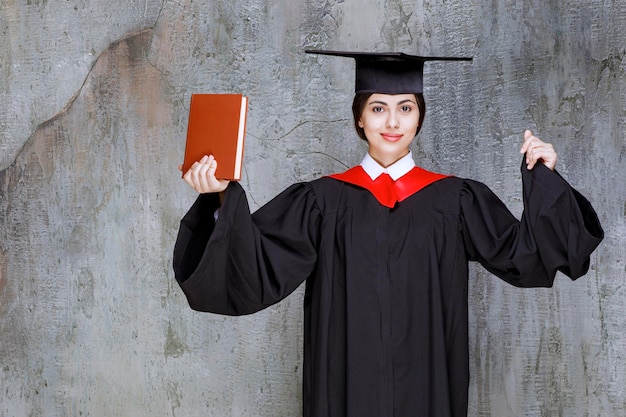 Portrait De Jeune étudiant Diplômé Avec Livre Debout Contre Le Mur. Photo De Haute Qualité