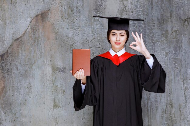 Portrait de jeune étudiant diplômé avec livre donnant le signe ok. Photo de haute qualité