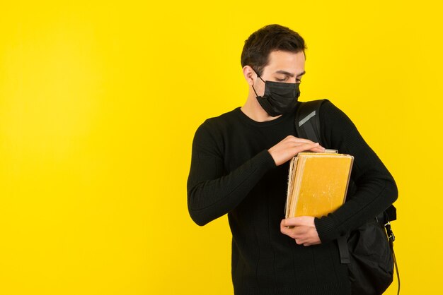 Portrait d'un jeune étudiant en masque médical tenant des livres d'université sur un mur jaune